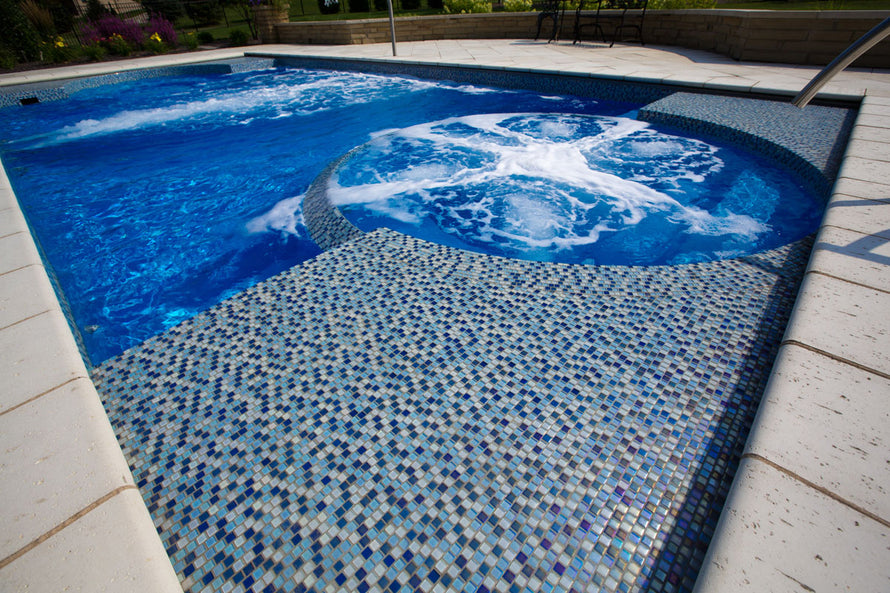 Image is a low angle view of a blue pool with blue tiles on the deck. Jets in the pool are aerating the water. 