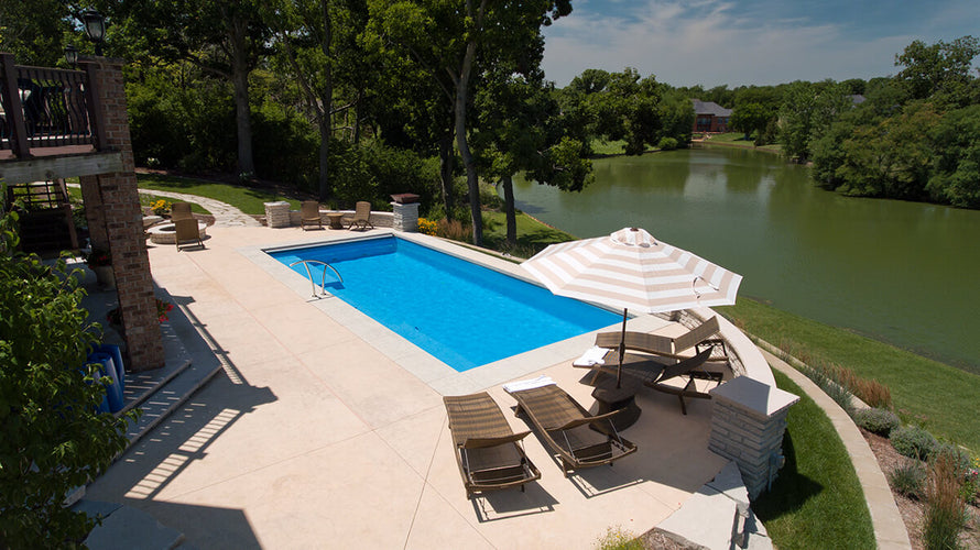View overlooking a patio. Chairs and umbrella set at the end of a crystal clear, rectangular pool.  Just to the right of the pool is the view of a lake. 